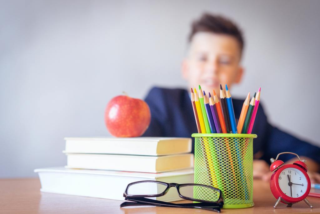 pencils in cup, clock, glasses, books, and apple in front of a student at a desk